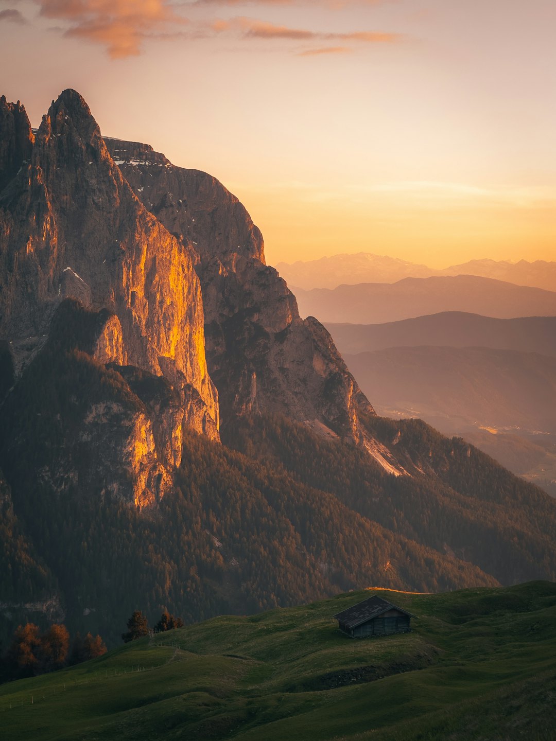 A small wooden cabin sits on a grassy hill before mountain peaks.